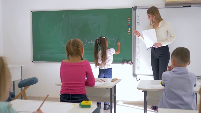 小学数学课上，女学生在教师旁边的黑板上写一个简单的例子视频素材