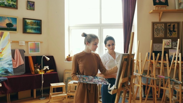 女美术老师正在与好看的年轻女孩学生绘画给建议站在画架前与画笔和调色板。视频素材