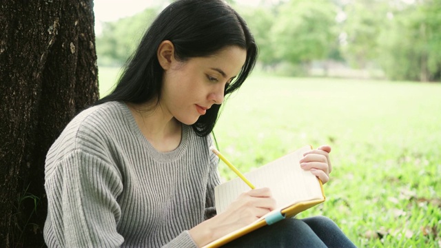 年轻迷人的女人在笔记本上写着什么，在公园里对着镜头微笑视频素材