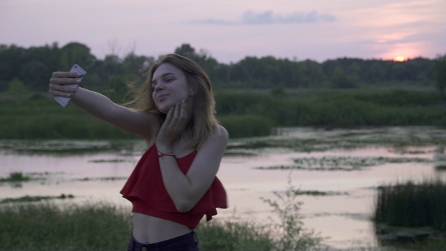 可爱的女孩摆拍手机在日落的背景上年轻的女人正在日落自拍女性正在户外休息视频素材