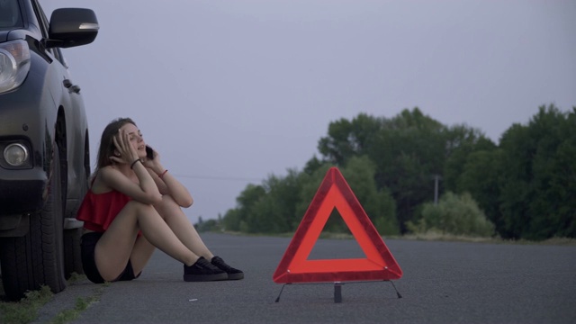 年轻女子坐在破车附近。女人是悲伤和孤独的。女性正在用手机打电话寻求帮助。女士的车出问题了视频素材