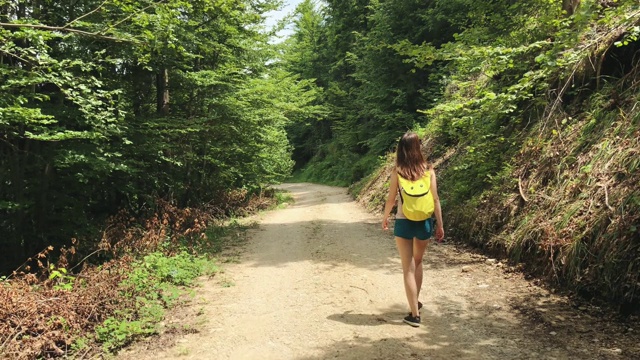 一个年轻的女性旅行者走在一个乡村的道路上与一个黄色的背包在野外的自然视频素材