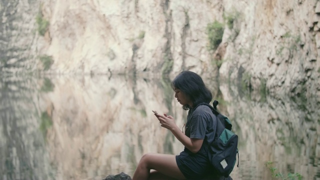 女登山者坐在河边，用智能手机拍摄周围美丽的风景。视频素材