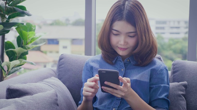 年轻的亚洲女子坐在家里的沙发上使用手机和微笑视频素材