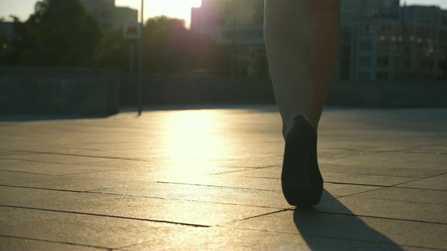 女性的腿在高跟鞋走在城市街道与太阳耀斑的背景。穿着高跟鞋的年轻女商人的脚在城市中穿行。女孩走着去上班。慢动作低角度视图视频素材