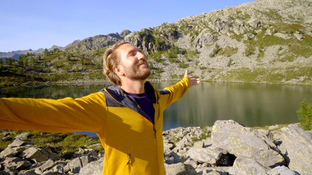 年轻男子徒步旅行在夏天通过惊人的高山湖泊手臂伸出庆祝个人目标。年轻人张开双臂，站在大自然中视频素材