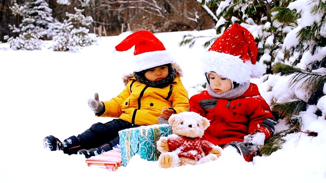 可爱的小男孩戴着圣诞帽，坐在雪地上放着新年礼物——库存视频视频下载