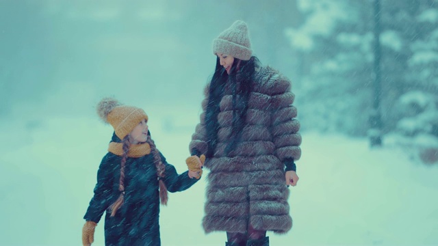 美丽的女人穿着裘皮大衣带着一个女孩在冬天里走在下雪的路上。他们正在散步。妈妈和女儿在谈笑。视频素材