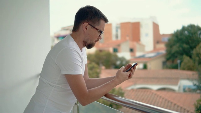 一名蓄着胡须的成年男子倚靠在阳台的扶手上看手机短信视频素材