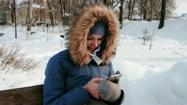 近景女子坐在长椅上浏览手机在冬天的公园里，在下雪的天气下，白天。视频素材