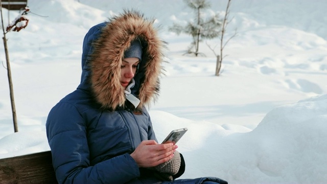 近景女子坐在长椅上浏览手机在冬天的公园里，在下雪的天气下，白天。视频素材