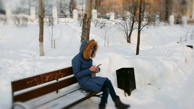 在大雪纷飞的冬日里，女子坐在城市公园的长椅上看手机视频素材