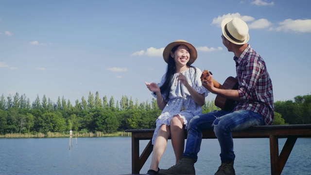 年轻的夫妇享受弹吉他，坐在湖边和蓝天的背景，复制空间视频素材