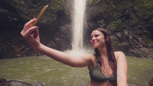 年轻女子旅游博主做自拍手机上的山瀑布背景。微笑的女人拍摄的手机自拍在飞溅的瀑布流动在雨林。视频素材