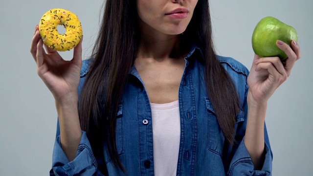 困惑的年轻女子拿着甜甜圈和苹果，营养选择，健康食品视频素材