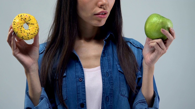 困惑的亚洲女人拿着甜甜圈和绿苹果，健康食品的选择，特写视频素材