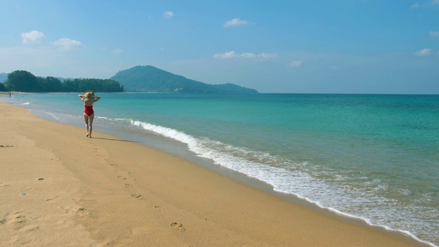 快乐微笑的年轻女子在太阳帽和红色泳衣沿着海边走在海滩上，夏天的海上度假和旅游概念视频素材