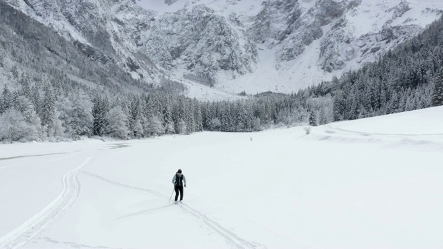 无人机拍摄的一名年轻女子在群山环绕的雪谷中越野滑雪视频素材