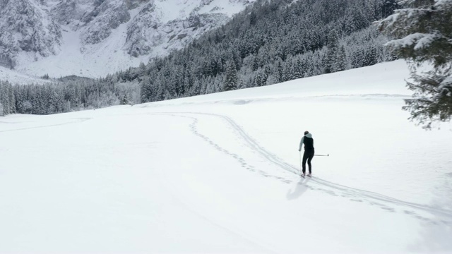 无人机拍摄的一名年轻女子在群山环绕的雪谷中越野滑雪视频素材