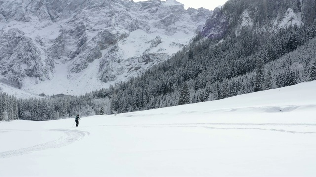 无人机拍摄的一名年轻女子在群山环绕的雪谷中越野滑雪视频素材