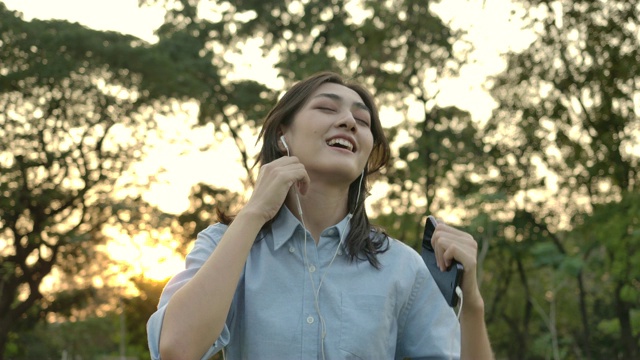 年轻迷人的亚洲女子听音乐手机和耳机享受声音舞蹈在夏季公园。有快乐情感观念的女人。缓慢的运动。视频素材