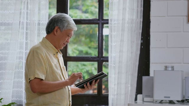 亚洲老年男性幸福地使用数字平板电脑，而站在家里的窗口，日常退休技术和生活方式视频素材