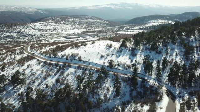 在恶劣天气下，俯视图无人机在弯曲的道路、风山和覆盖着积雪的森林上空飞行。视频素材