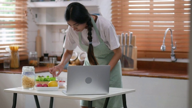 年轻的亚洲女子用笔记本电脑和配料做沙拉，蔬菜，草莓在家里厨房做一份健康的早餐。视频素材