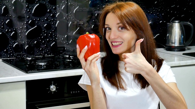 有吸引力的兴奋的年轻女子与大红色柿子椒显示拇指坐在厨房在她的家视频素材