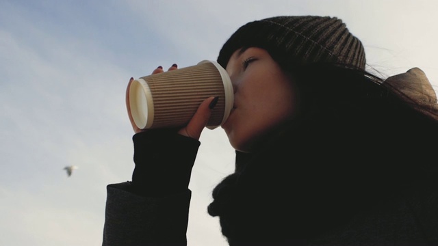 戴着帽子和围巾的女孩在海边用一次性杯子喝茶视频素材