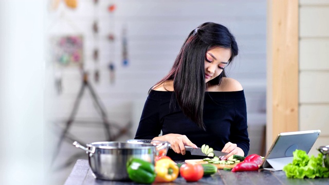 中镜头漂亮微笑的年轻亚洲女人坐在厨房的桌子上切黄瓜视频素材