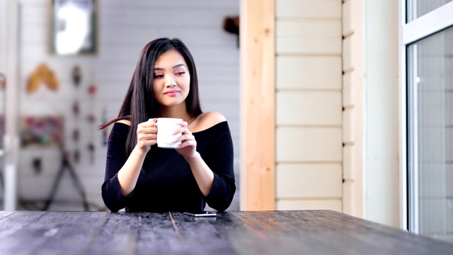 迷人的年轻亚洲女人喝热咖啡或茶坐在桌子上看着窗户视频素材