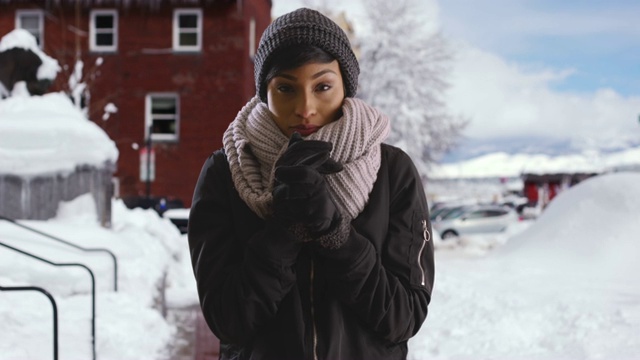 年轻的黑人妇女站在白雪覆盖的街道上，向手中吹气取暖视频素材