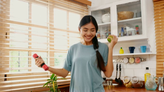 年轻的亚洲妇女拿着哑铃和青苹果在厨房里锻炼，人们和健康的生活方式视频素材