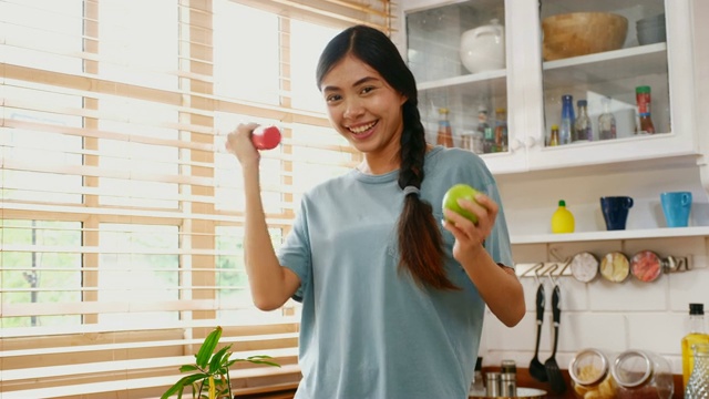 年轻的亚洲妇女拿着哑铃和青苹果在厨房里锻炼，人们和健康的生活方式视频素材