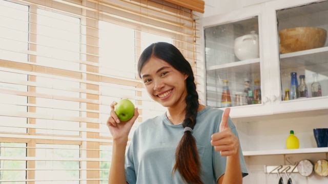 年轻的亚洲女人拿着苹果微笑和拇指与幸福在厨房在家里，人们健康的生活方式视频素材