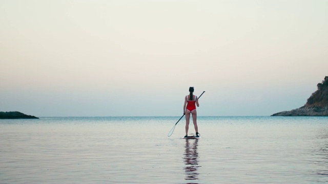 女人在红色泳衣上SUP站立桨板在海湾日落视频素材