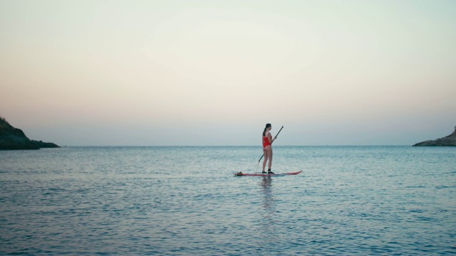 女人在红色泳衣上SUP站立桨板在海湾日落视频素材