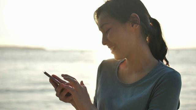 亚洲女人在海滩上的日落使用智能手机视频素材