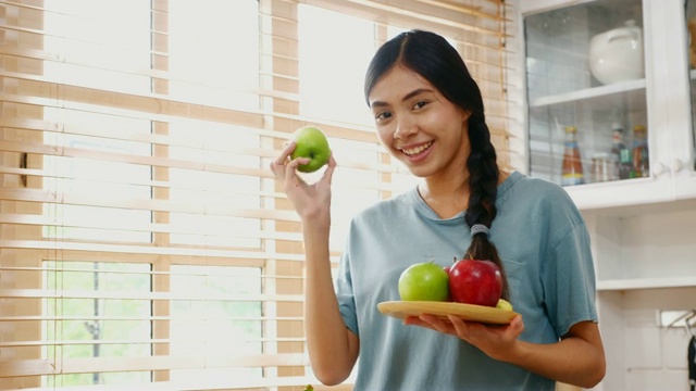 年轻的亚洲女人拿着苹果与幸福在家里的厨房，人们健康的生活方式视频素材