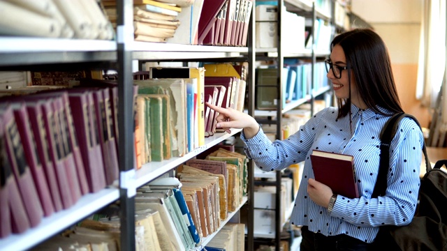 年轻的学生拿着书和背包站在大学图书馆的书架旁视频素材