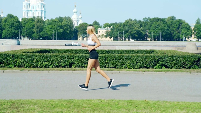 跟踪左图的fit blonde女人在柏油路上跑过绿色的夏天公园旁边的河流在慢动作视频素材