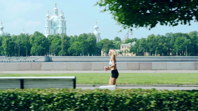 在温暖的夏日里，年轻健康的女人穿着运动胸罩和短裤跑过河边和教堂附近的公园视频素材