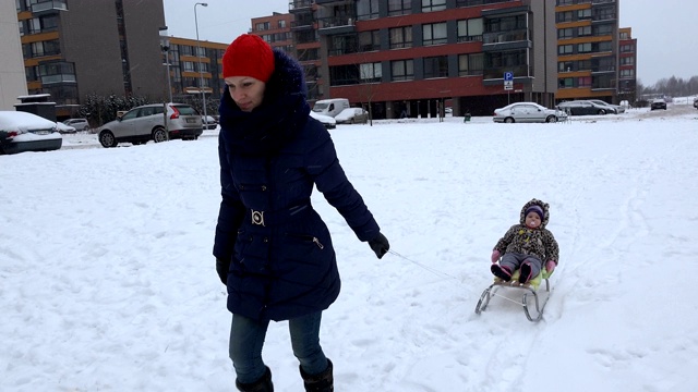 在居住区，母亲拉着雪橇在雪地上拉着女儿的婴儿。4 k视频素材