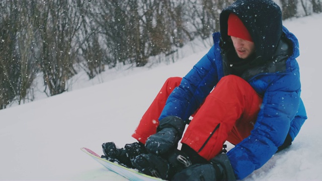 一个穿着红色裤子的男人坐在雪地上，在滑雪坡上系着滑雪板靴视频素材