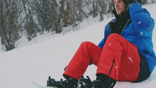 一个穿着红色裤子的男人坐在雪地上系着滑雪板鞋和蓝色夹克在滑雪坡上视频素材