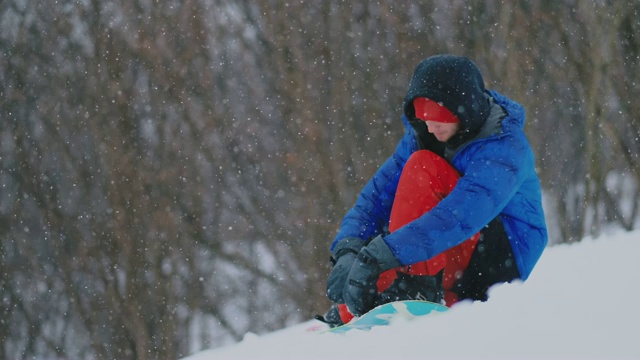 一个穿着红色裤子的男人坐在雪地上系着滑雪板鞋和蓝色夹克在滑雪坡上视频素材