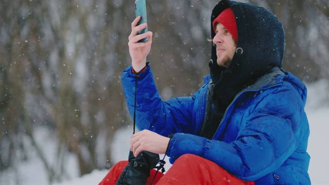 男滑雪板坐在雪地上用手机拍摄度假村的美丽风景，用于社交网络视频素材