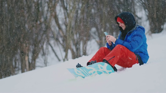 一名男性滑雪者坐在雪地上用手机拍摄一处美丽的度假胜地风景，用于社交网络。度假村的博主。用你的智能手机给朋友发短信视频素材