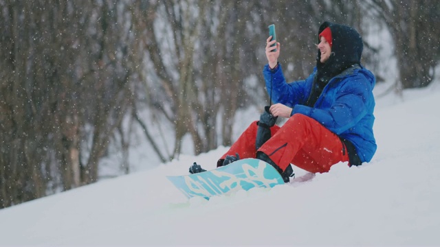 男滑雪板坐在雪地上用手机拍摄度假村的美丽风景，用于社交网络视频素材